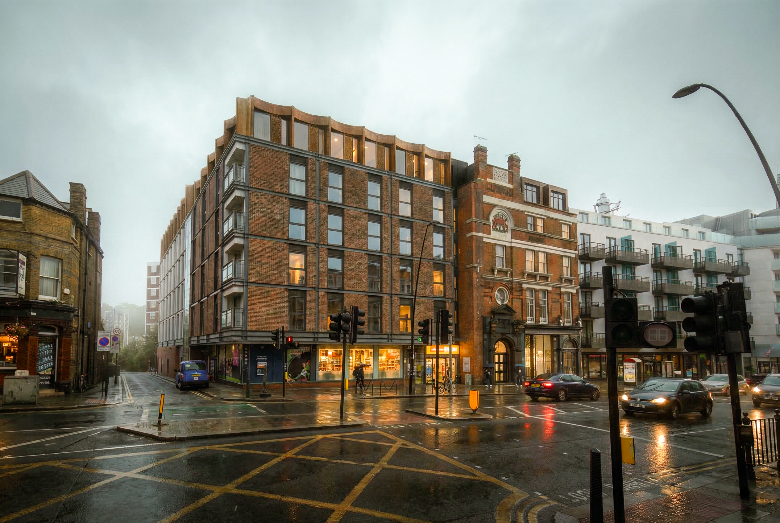Dusk CGI on a rainy evening of the proposed hotel at 34 and 34A–36 Kilburn High Road — the refurbished brick buildings with new single-storey rooftop extensions and warm-lit windows, alongside the retained historic frontage of the former Red Lion, reflected on the wet street.